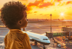 Children's Passport Requirements in the USA - A young Black boy with curly hair, wearing a mustard-colored shirt, gazes thoughtfully out of an airport window as the golden hues of a sunset light up the sky. In the foreground, a plane is parked at the gate, with the airport runway and additional aircraft visible in the background. The scene captures a moment of anticipation and wonder, evoking the excitement of travel and exploration